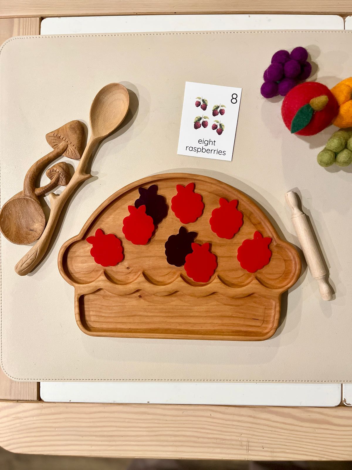 Wooden pie-shaped tray with seven red raspberry-shaped pieces, wooden utensils, felt fruit, and an educational card nearby