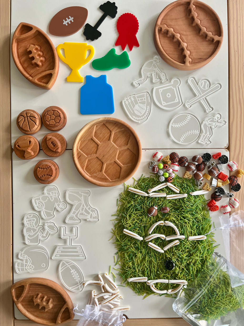 Assortment of wooden and plastic sports-themed shapes and cookie cutters arranged on a white surface with scattered green grass-like pieces
