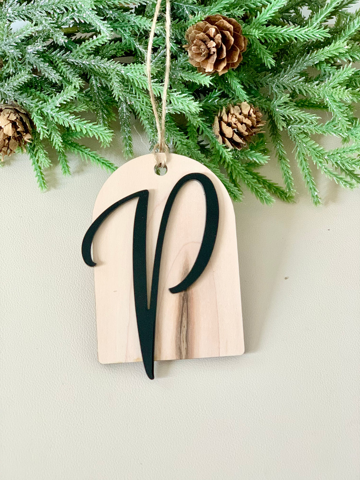Light natural wood arch ornament with a black cursive letter V hanging from twine against pine branches and cones