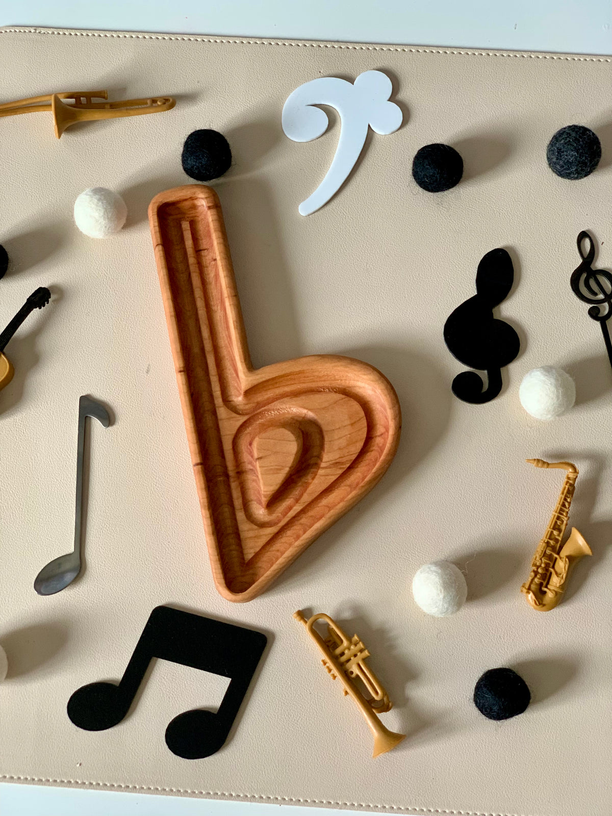 Wooden bass clef tray surrounded by black and white felt balls, small musical notes, and miniature brass instruments on beige surface