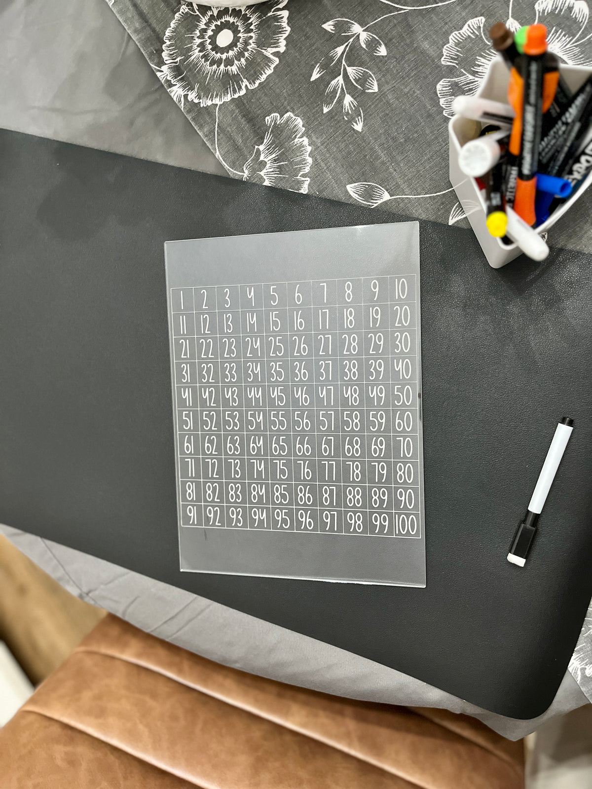 Clear acrylic sheet with numbered grid 1 to 100 beside a black dry erase marker and container of colorful markers on table