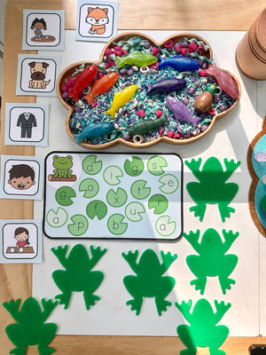 Cloud-shaped wooden sensory tray filled with colorful fish toys and dyed rice, surrounded by green frog mats and letter cards on wooden table