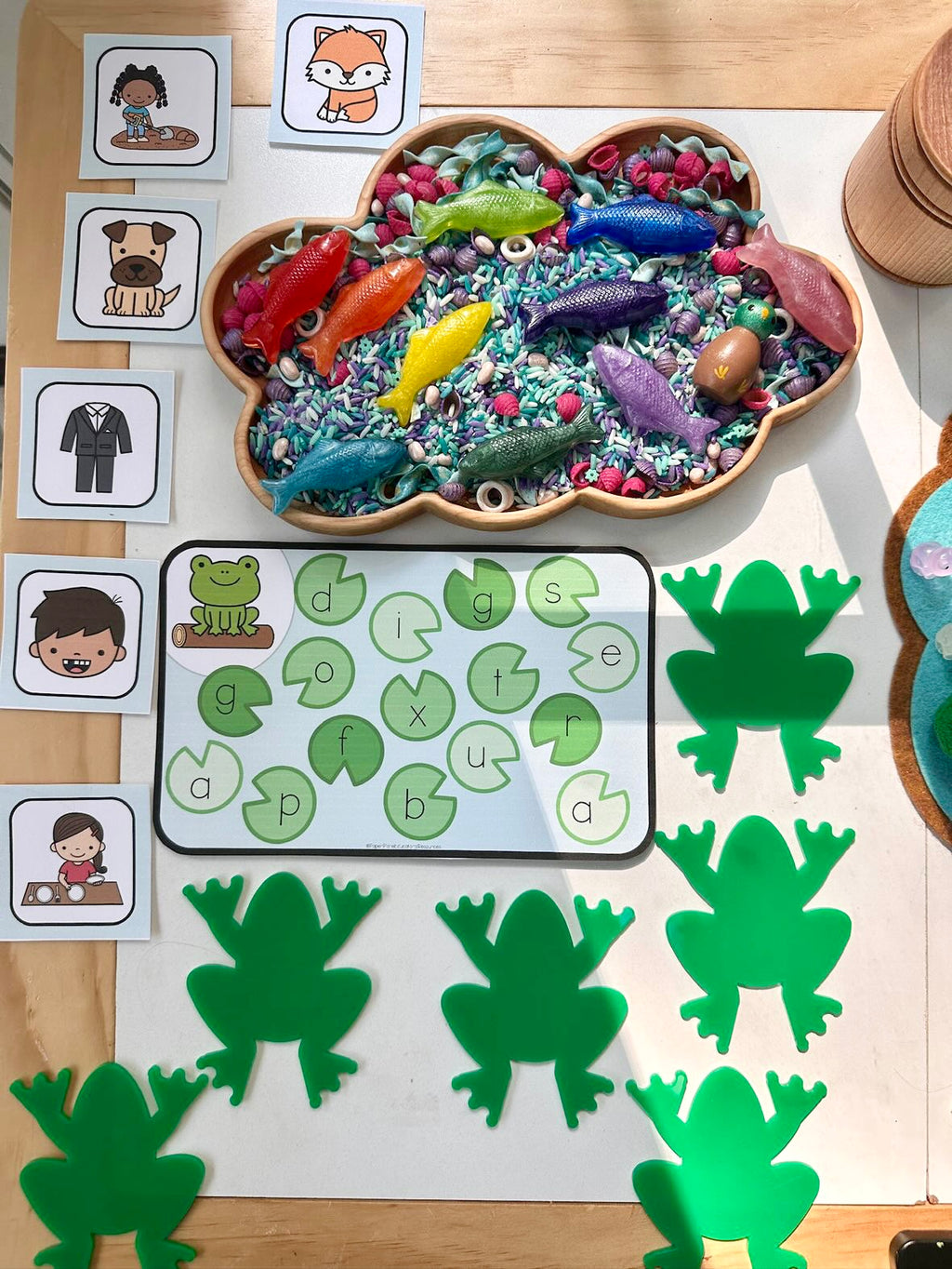 Cloud-shaped wooden sensory tray filled with colorful fish toys and dyed rice, surrounded by green frog mats and letter cards on wooden table