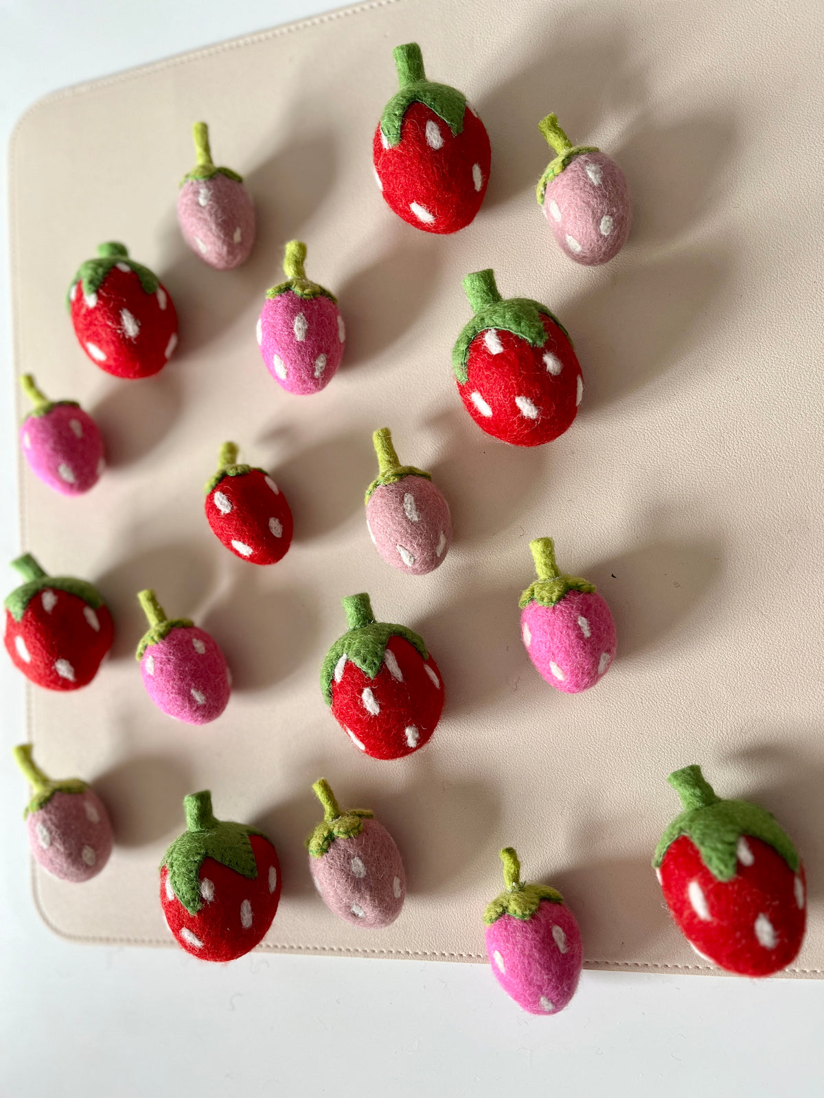 Assorted small felt strawberries in red, pink, and purple shades arranged on a light beige surface
