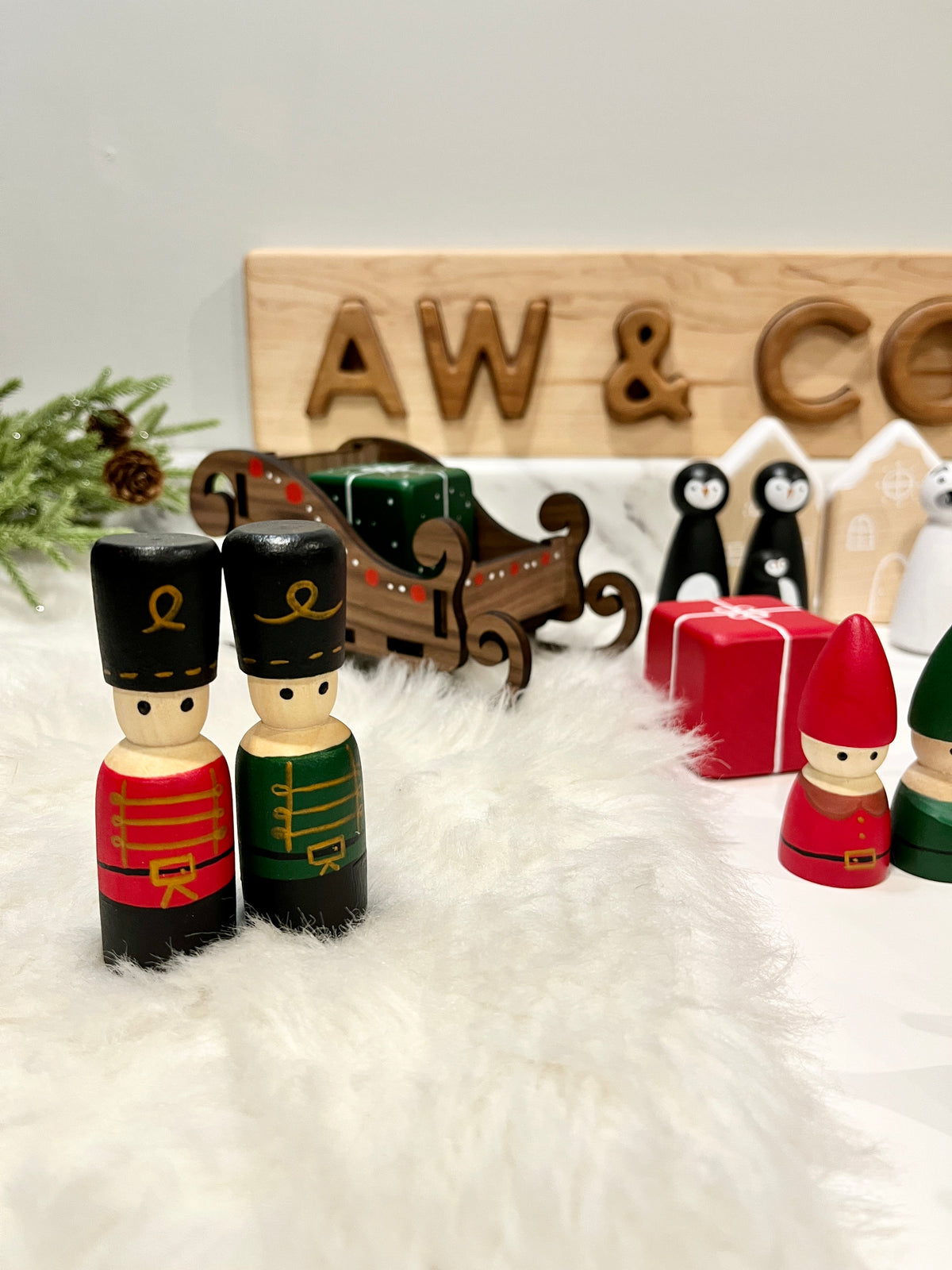 Wooden toy soldiers, sleigh, gift box, gnome figures, and penguin ornaments arranged on white fur with greenery background