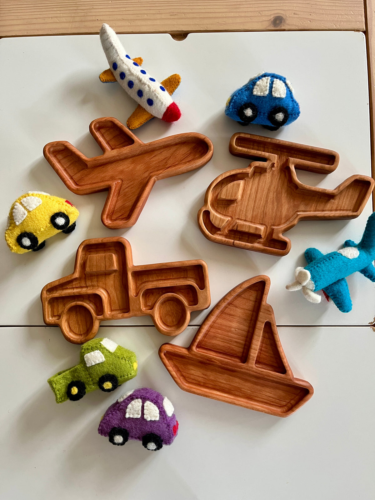 Wooden trays shaped like airplane, helicopter, boat, and truck surrounded by colorful felt toy vehicles on table