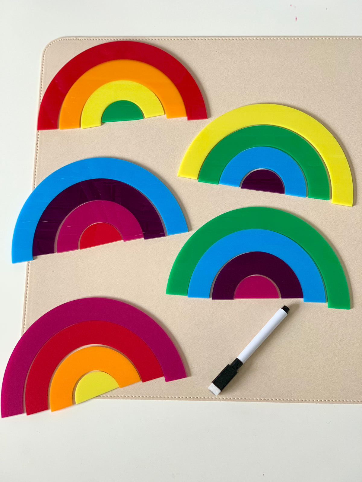 Five colorful plastic rainbow-shaped stacking pieces arranged on a beige surface with a black and white marker nearby
