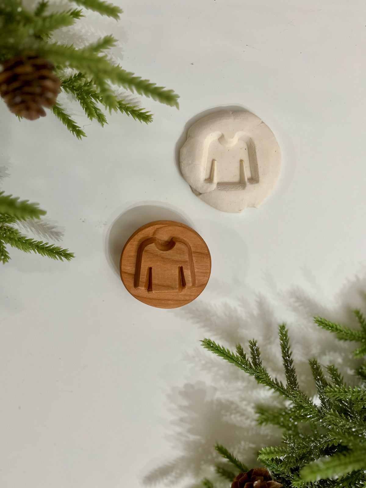 Round wooden cookie stamp with sweater design and matching white dough impression on a white surface with green pine branches