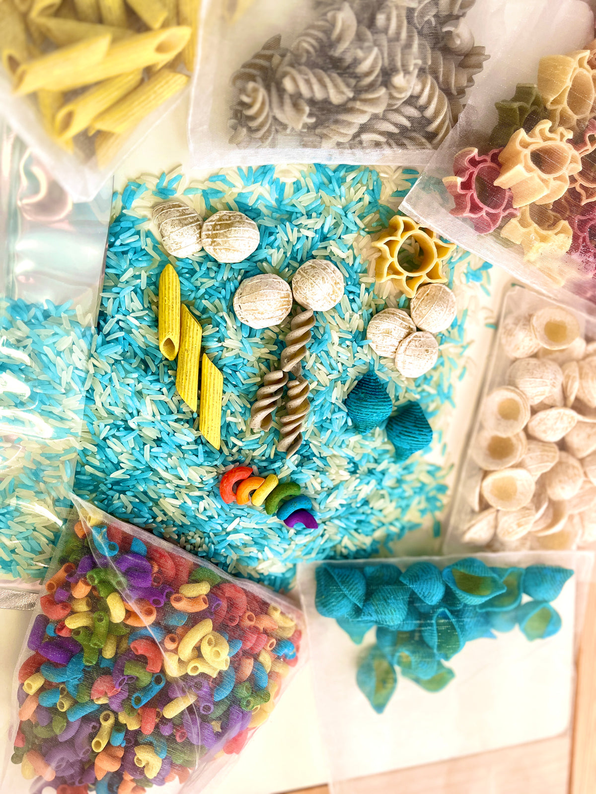 Various colorful pasta shapes and textured items arranged on blue and white rice background with clear bags nearby