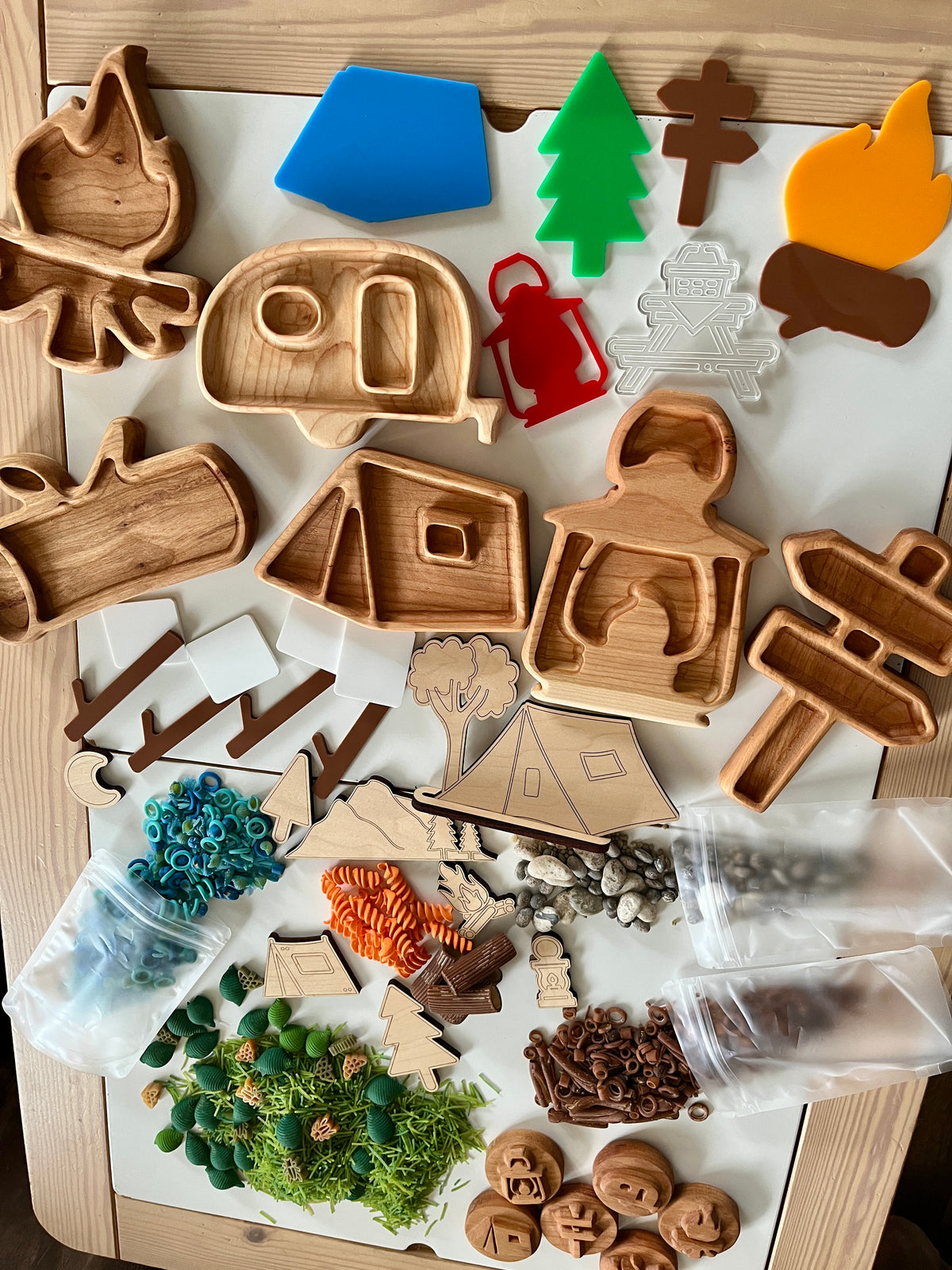 Wooden camping-themed trays, colorful camping icons, faux foliage, rocks, and small toy accessories on a light wooden surface
