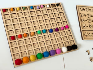 Wooden laser cut hundred board sensory tray with colorful felt balls in 10 rows on white table with wooden number puzzle nearby