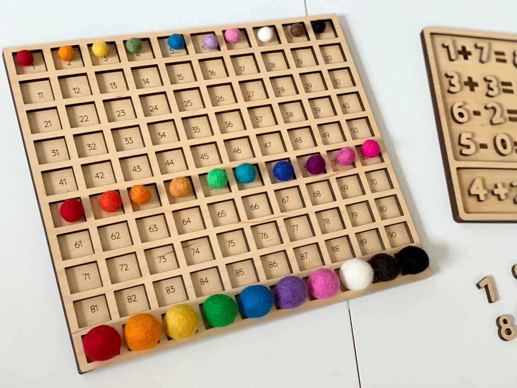 Wooden laser cut hundred board sensory tray with colorful felt balls in 10 rows on white table with wooden number puzzle nearby
