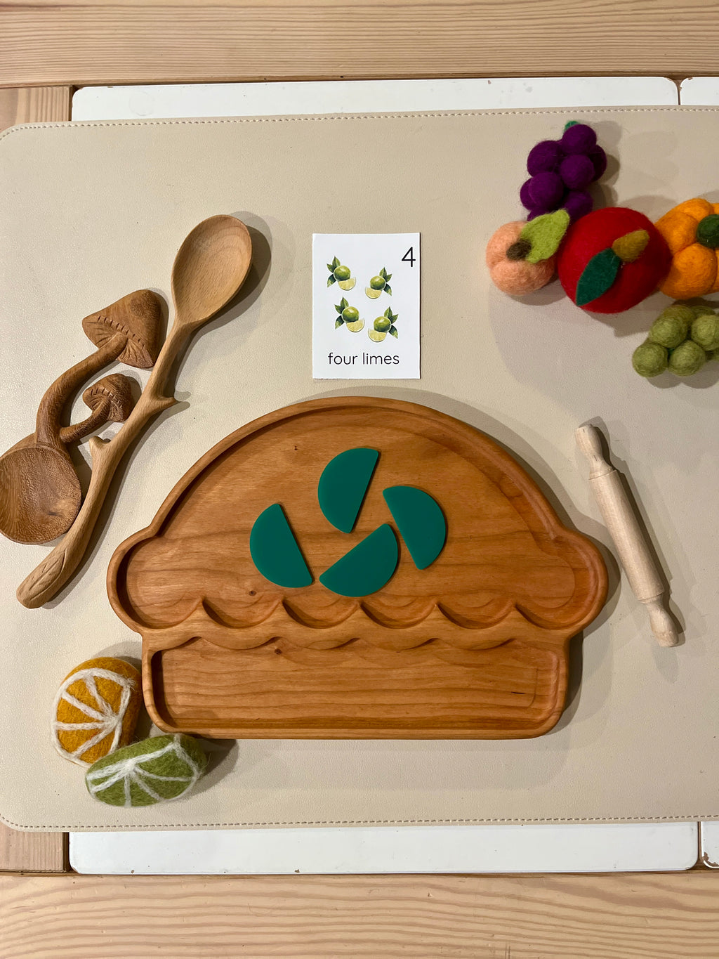 Wooden pie-shaped tray with four green felt lime slices, wooden utensils, rolling pin, felt fruit, and a flashcard on beige surface