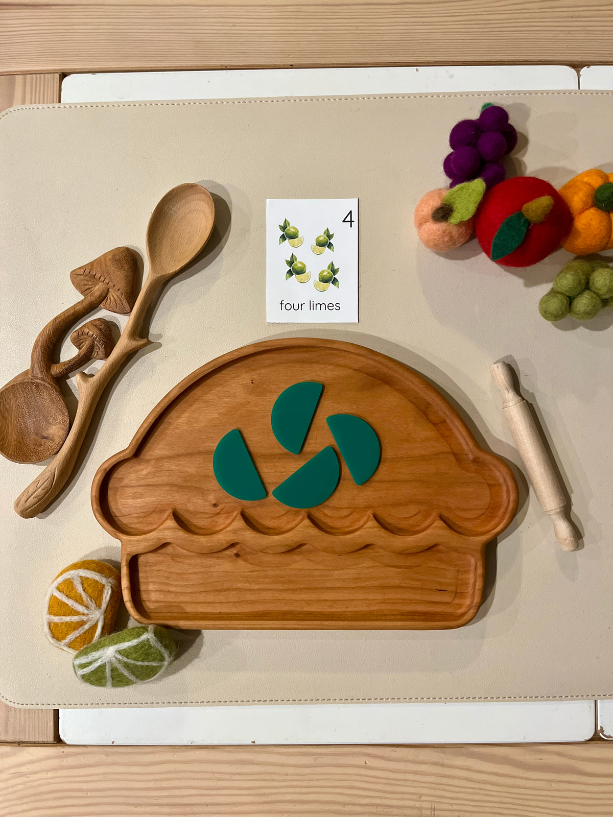 Wooden pie-shaped tray with four green felt lime slices, wooden utensils, rolling pin, felt fruit, and a flashcard on beige surface