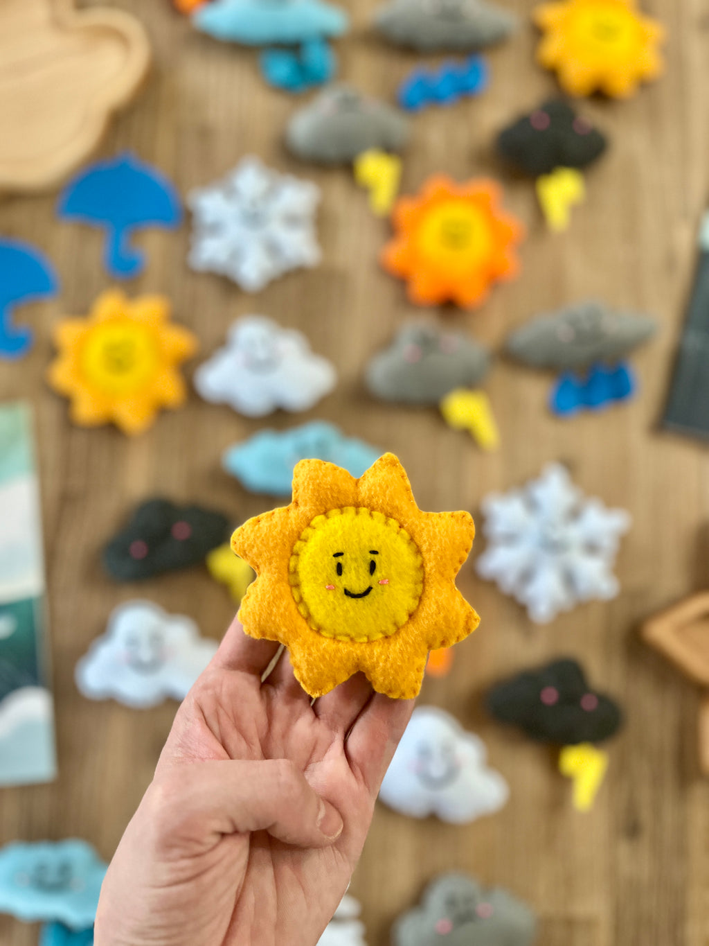 Hand holding a smiling orange felt sun with other colorful felt weather shapes including clouds, snowflakes, and lightning in the background, Felt Weather Shapes, Made in Canada, handcrafted, homeschool, education resources.