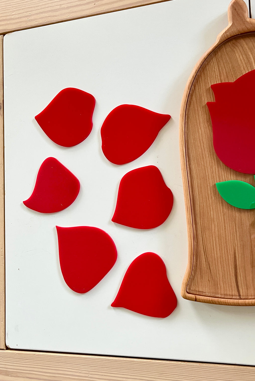 Six red acrylic pieces shaped like varied flower petals arranged next to a wooden tray with a partial floral design