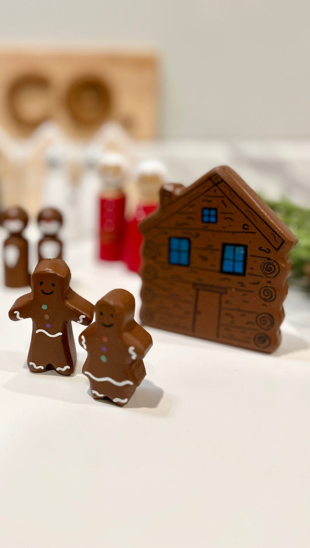 Wooden gingerbread man and woman figurines with a carved log cabin and small figurines blurred in the background