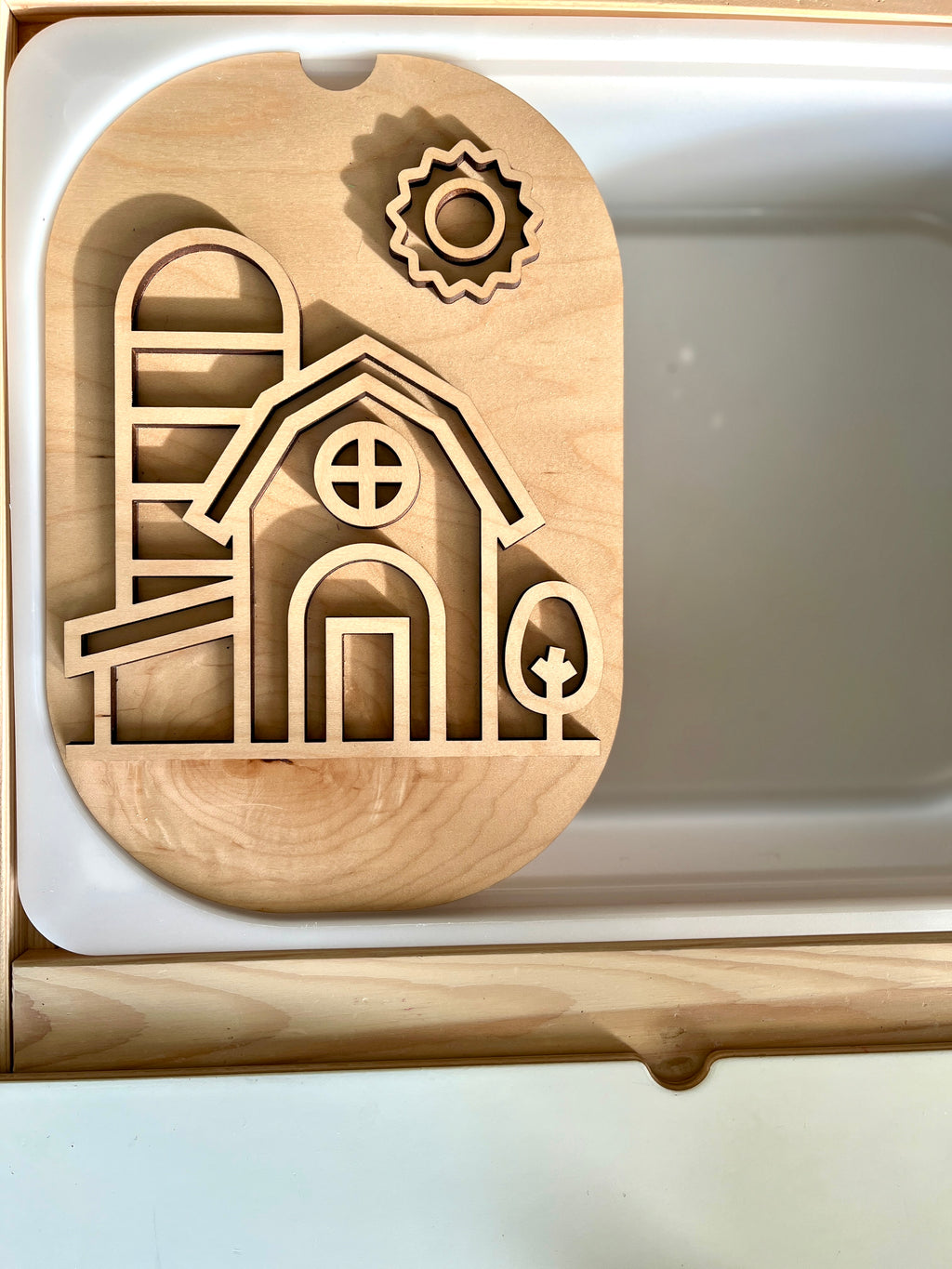 Light wooden cutout board depicting a barn, silo, sun, and tree shapes resting inside a white compartment tray.