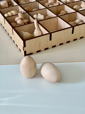 Two smooth unfinished wooden egg shapes placed in front of a divided wooden storage box with various wooden pieces inside