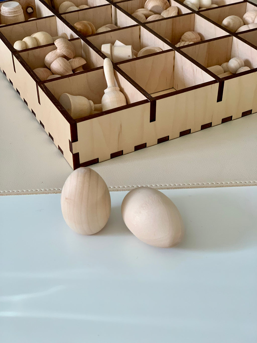Two smooth unfinished wooden egg shapes placed in front of a divided wooden storage box with various wooden pieces inside