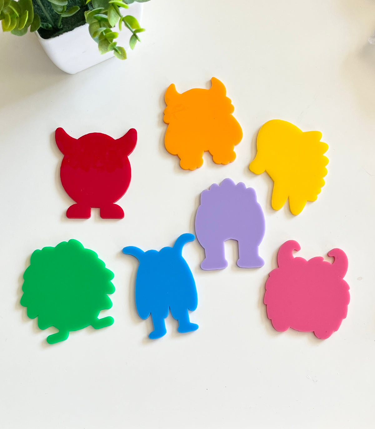 Seven colorful monster-shaped cutouts arranged on a white surface near a potted green plant