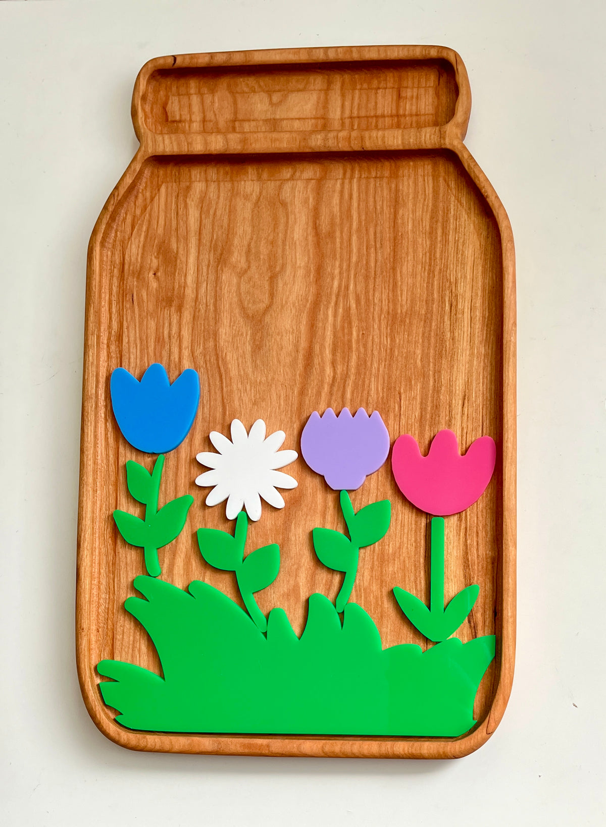 Wooden tray shaped like a jar with colorful plastic flowers and green leaves arranged at the bottom center