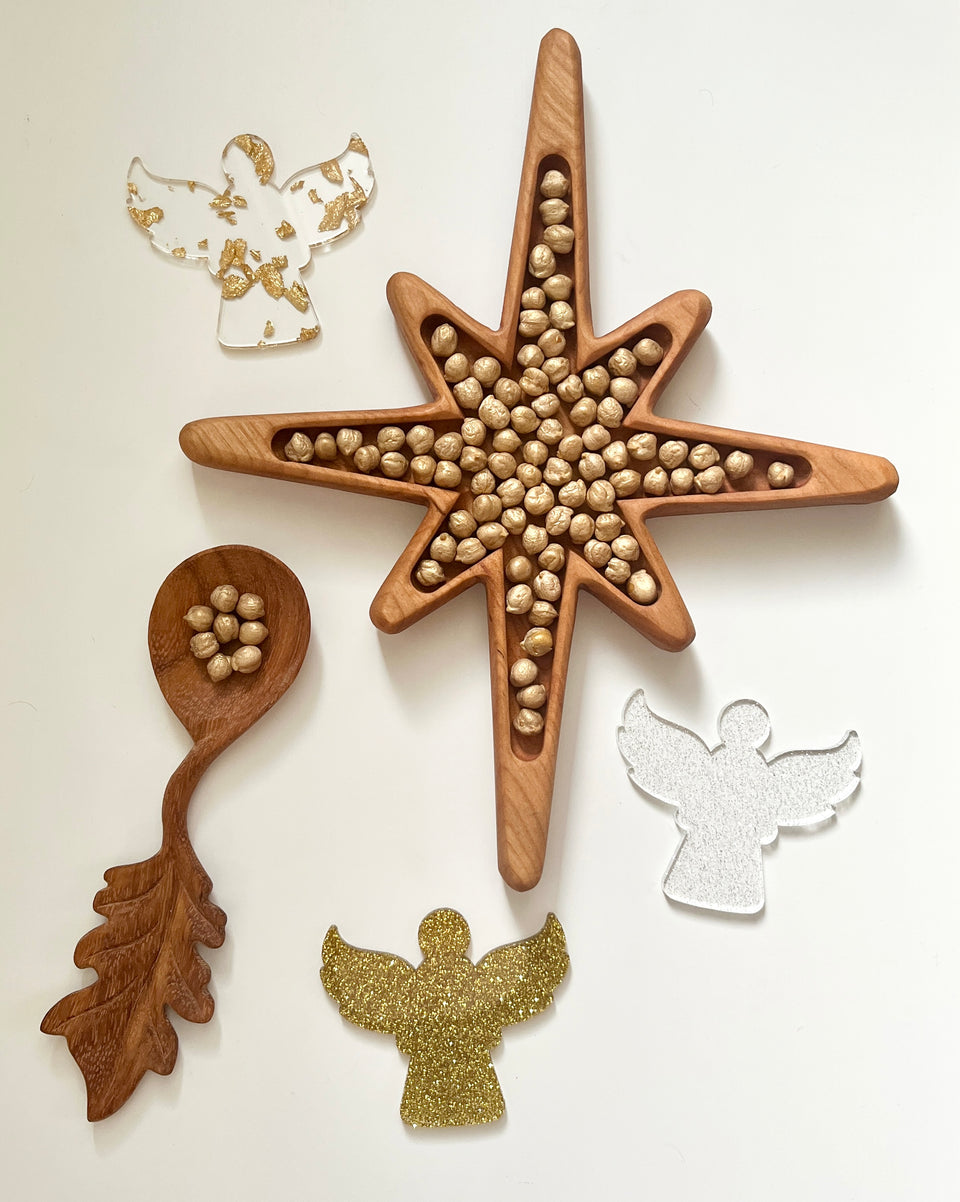 Star-shaped wooden tray filled with hazelnuts next to wooden spoon and three decorative angel cutouts