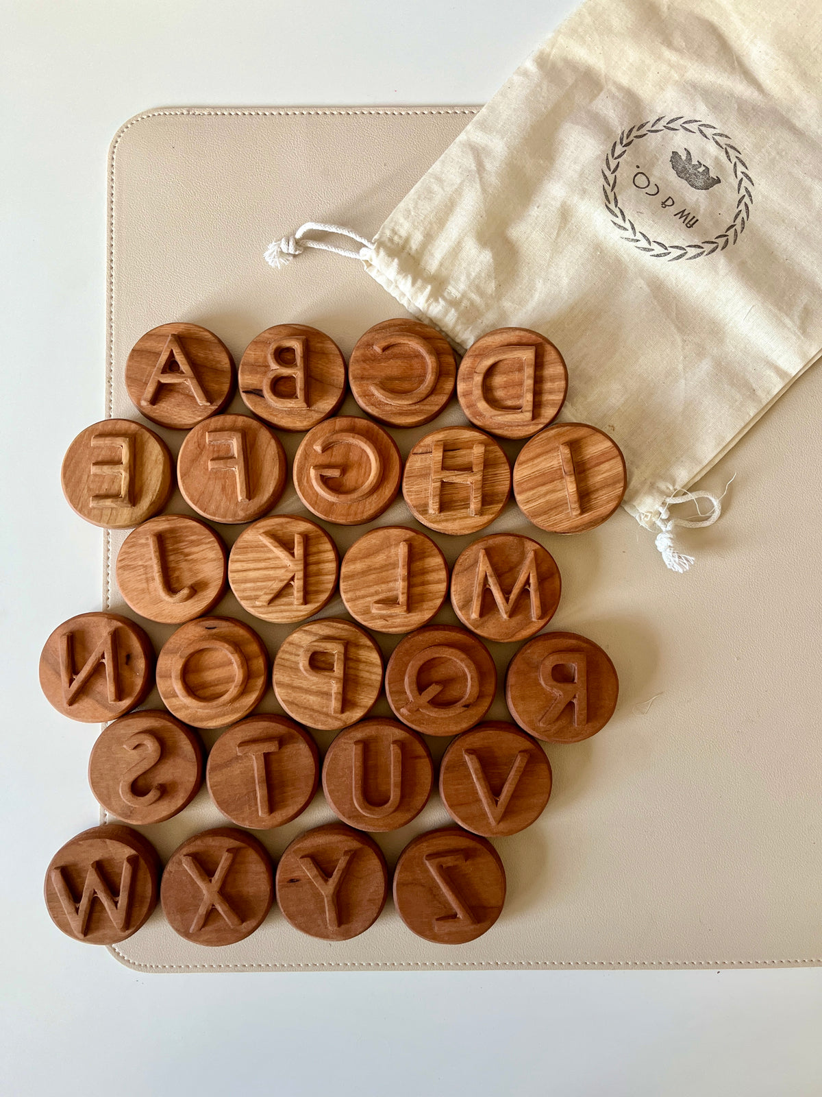 Set of round wooden letter stamps with uppercase alphabet characters next to a beige drawstring storage bag on a white surface