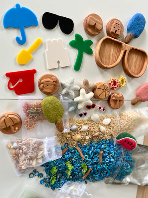 Collection of colorful beach-themed tokens, wooden trays, felt popsicles, shells, seeds, and sensory play materials on a white surface