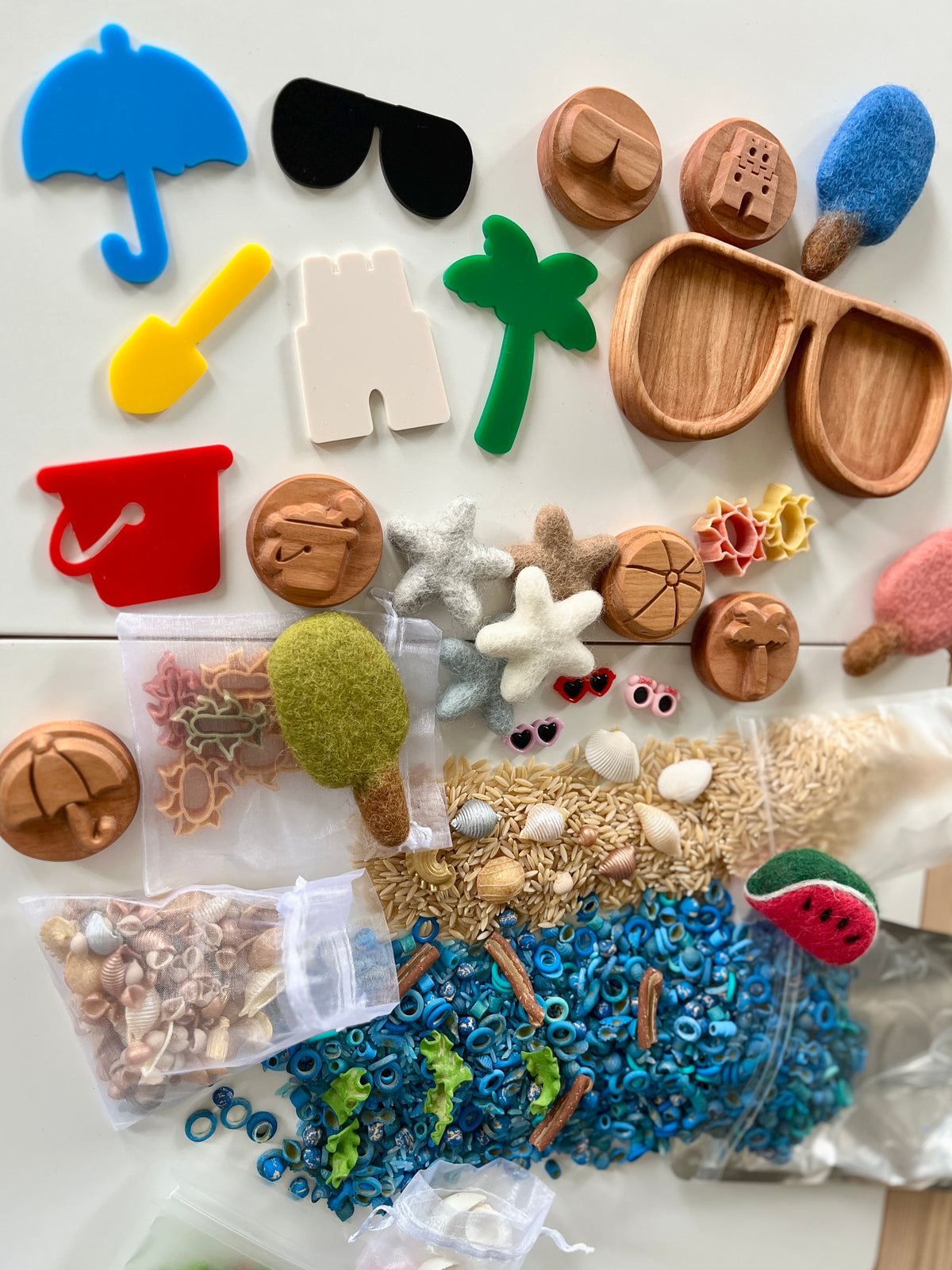 Collection of colorful beach-themed tokens, wooden trays, felt popsicles, shells, seeds, and sensory play materials on a white surface