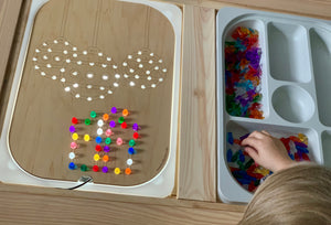 Wooden Flisat insert with holes for colorful pegs forming Christmas ball and gift shapes beside trays of translucent pegs and child's hand