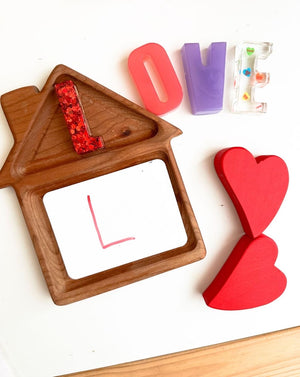 Natural wooden house-shaped sensory tray with colorful resin letter pieces and red wooden hearts on white surface