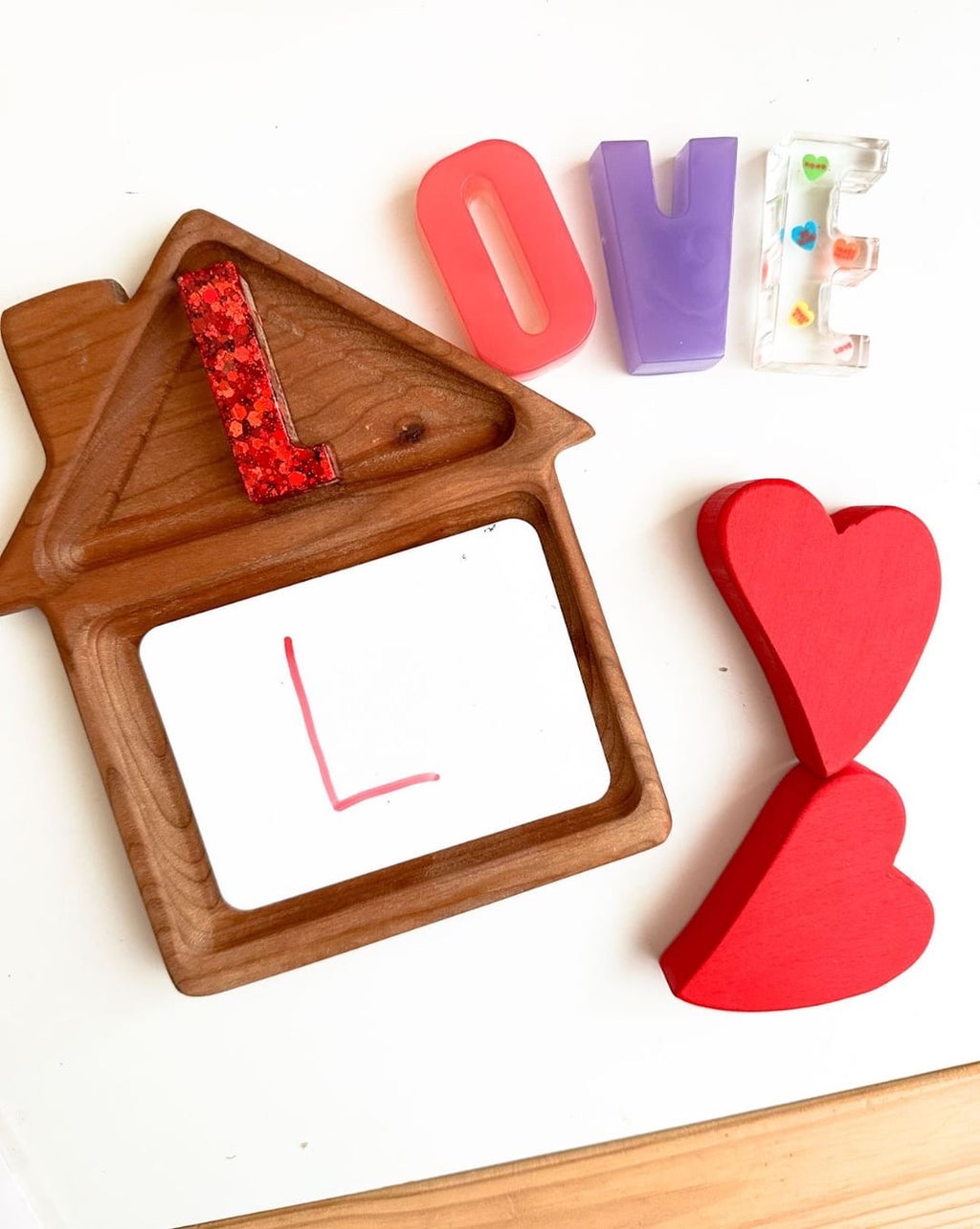 Natural wooden house-shaped sensory tray with colorful resin letter pieces and red wooden hearts on white surface