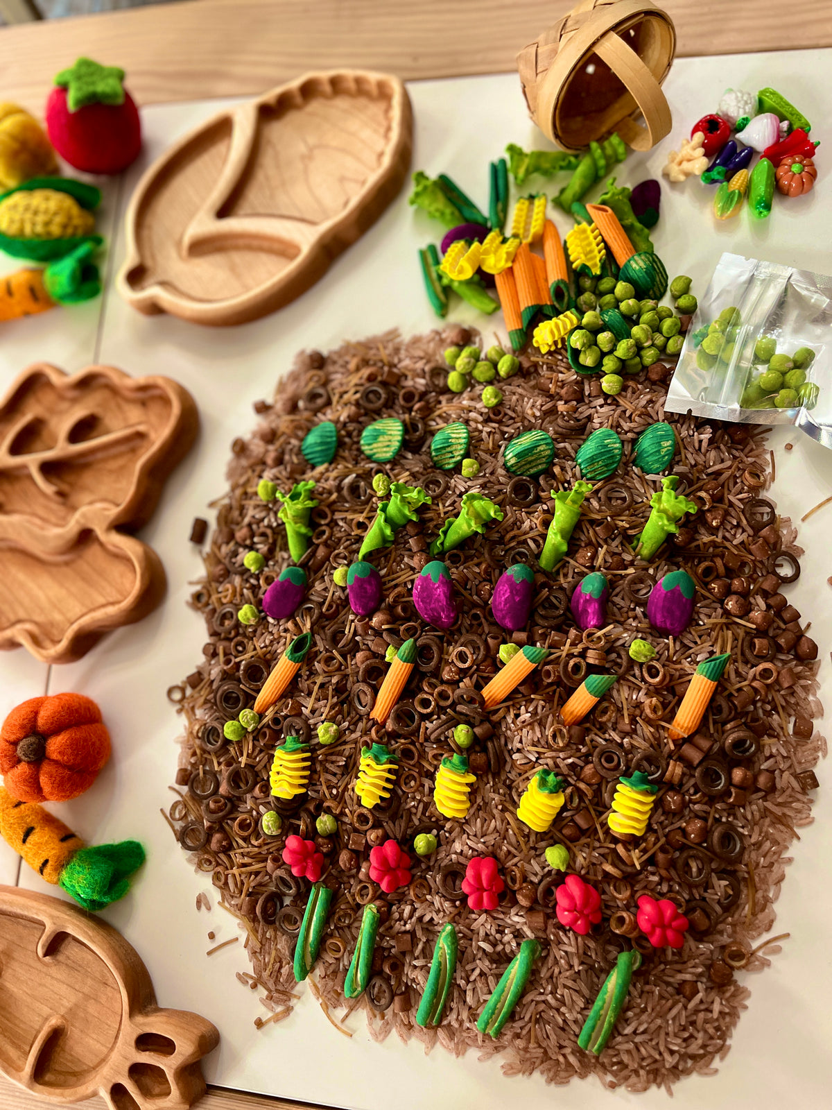Miniature clay vegetables and pasta arranged artfully on brown rice with wooden trays and felt produce nearby