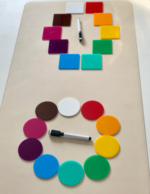 Colorful square and circular acrylic tiles arranged in circles with two black and white dry erase markers on a beige surface
