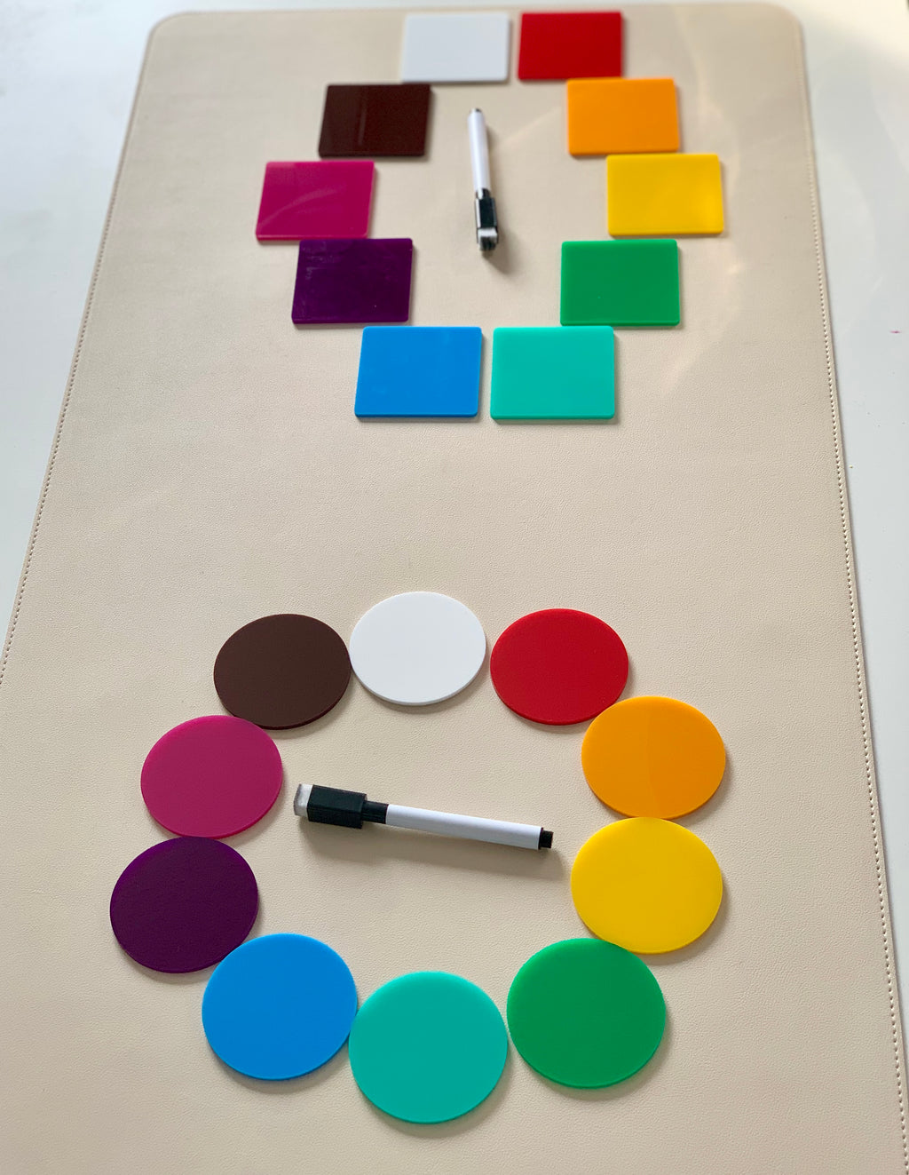 Colorful square and circular acrylic tiles arranged in circles with two black and white dry erase markers on a beige surface
