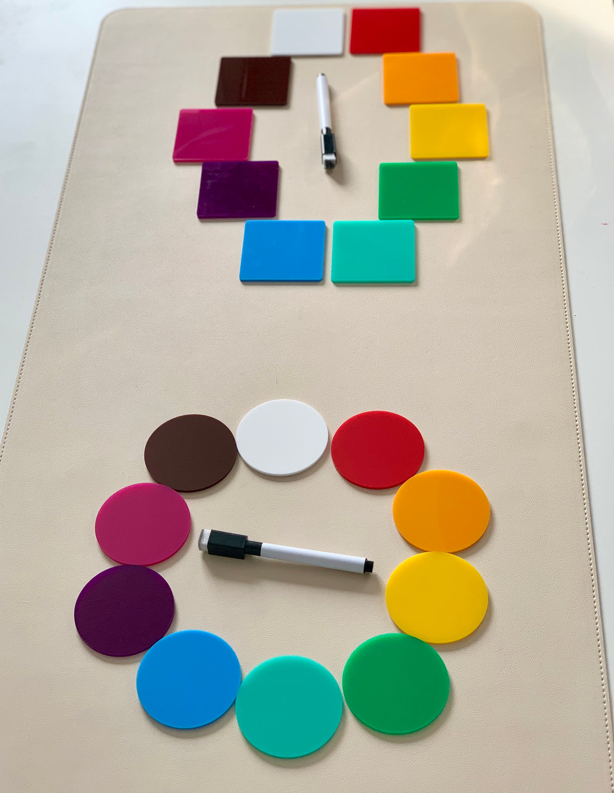 Colorful square and circular acrylic tiles arranged in circles with two black and white dry erase markers on a beige surface
