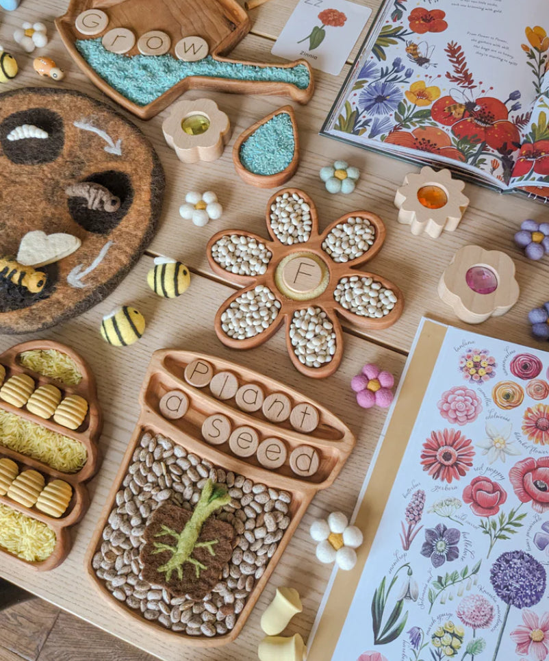 Wooden plant and flower-shaped trays filled with seeds, felt bees, flower decor, and a botanical guide on a wood table