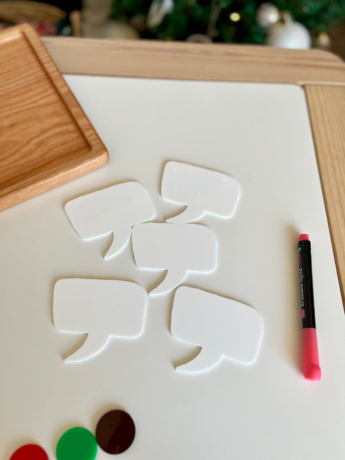 Five blank white speech bubble cutouts and a pink marker on a white board with a wooden tray nearby