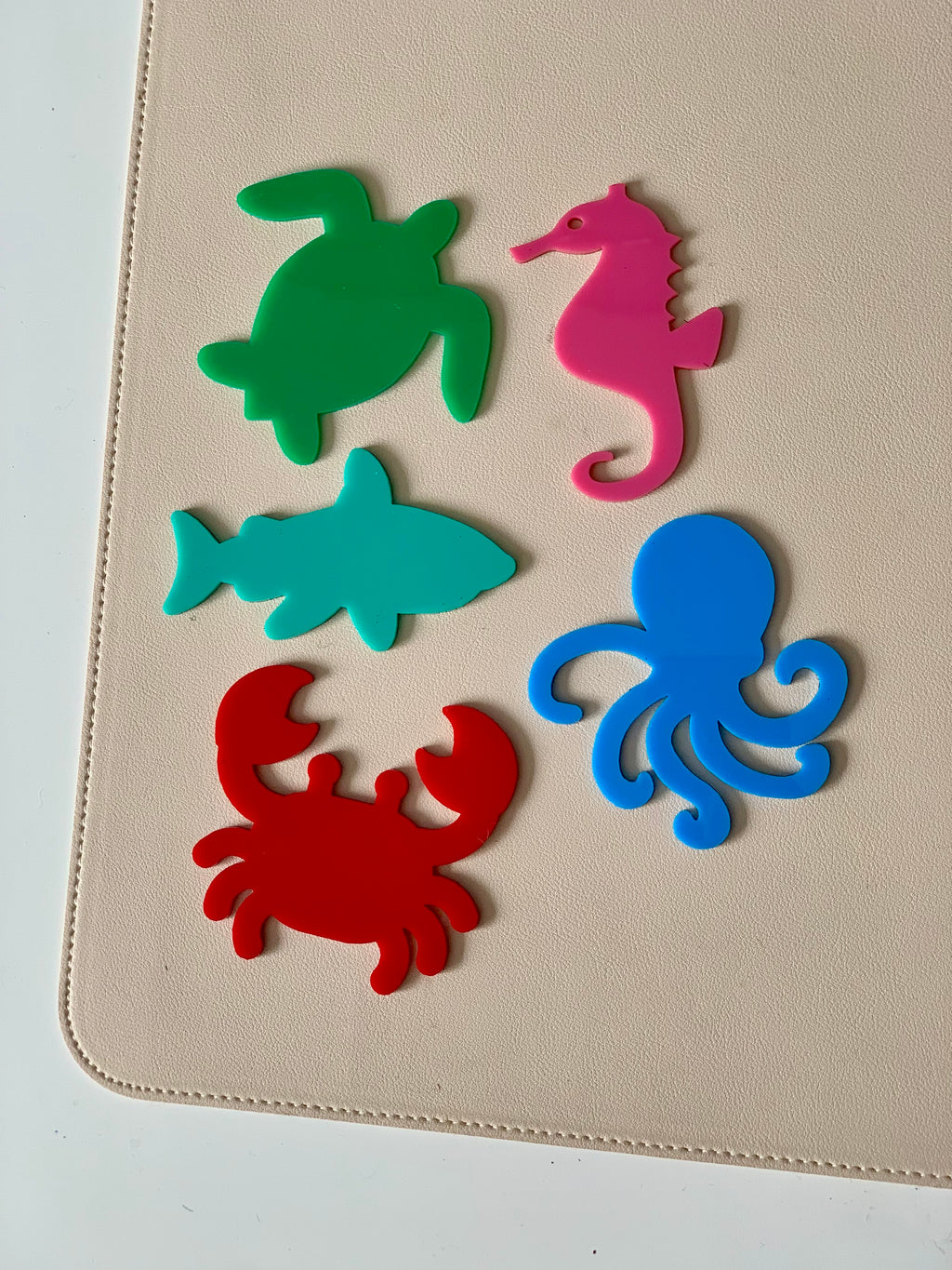 Five colorful plastic sea creature cutouts including a green turtle, pink seahorse, teal shark, red crab, and blue octopus on beige surface