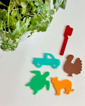 Five colorful acrylic shapes including red mailbox, turquoise truck, brown turkey, green turtle, and orange cat on white surface with green leaves above