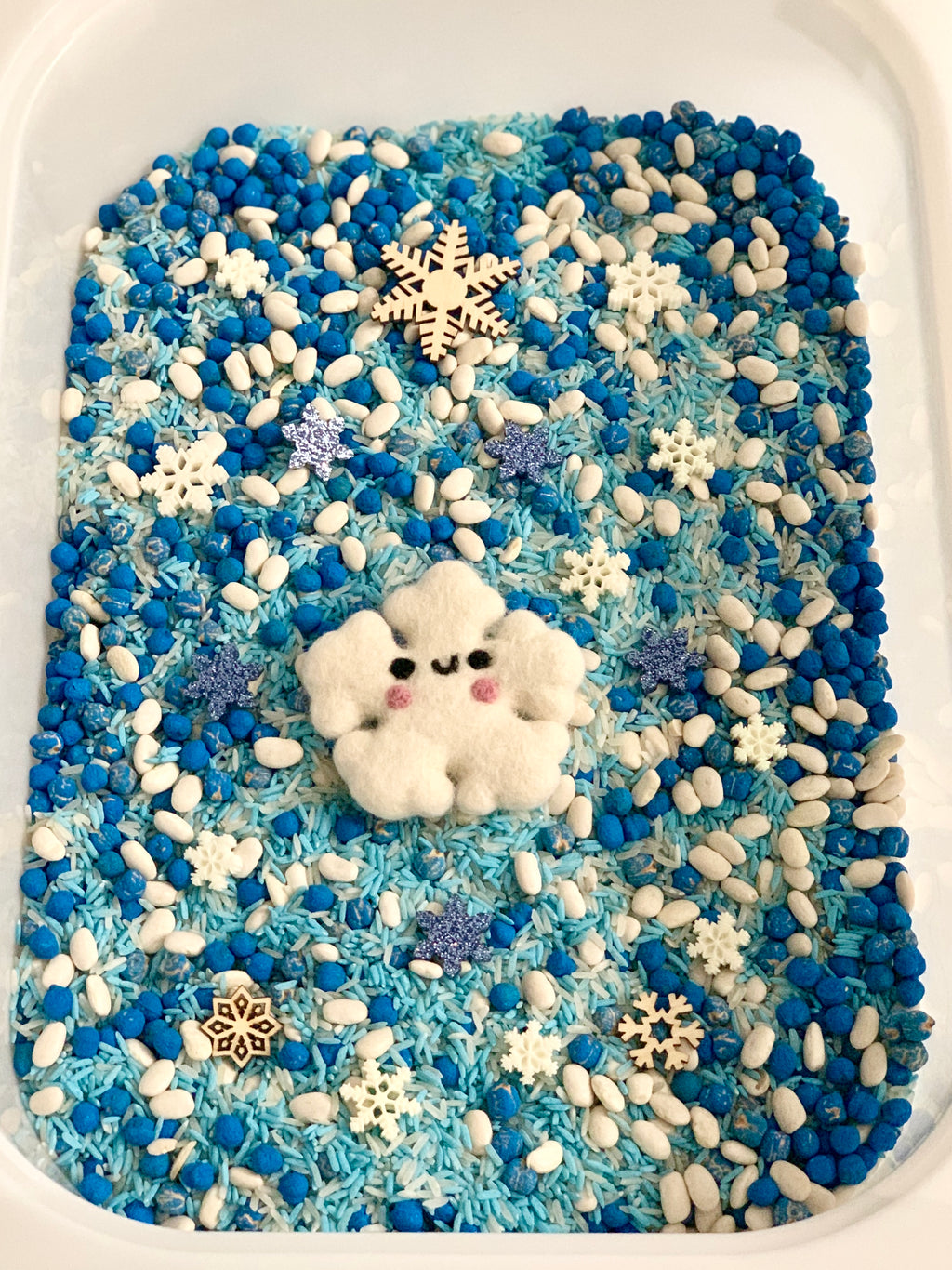 Blue Christmas sensory bin filled with blue and white rice, beans, snowflake shapes, and a smiling felt snowflake centerpiece