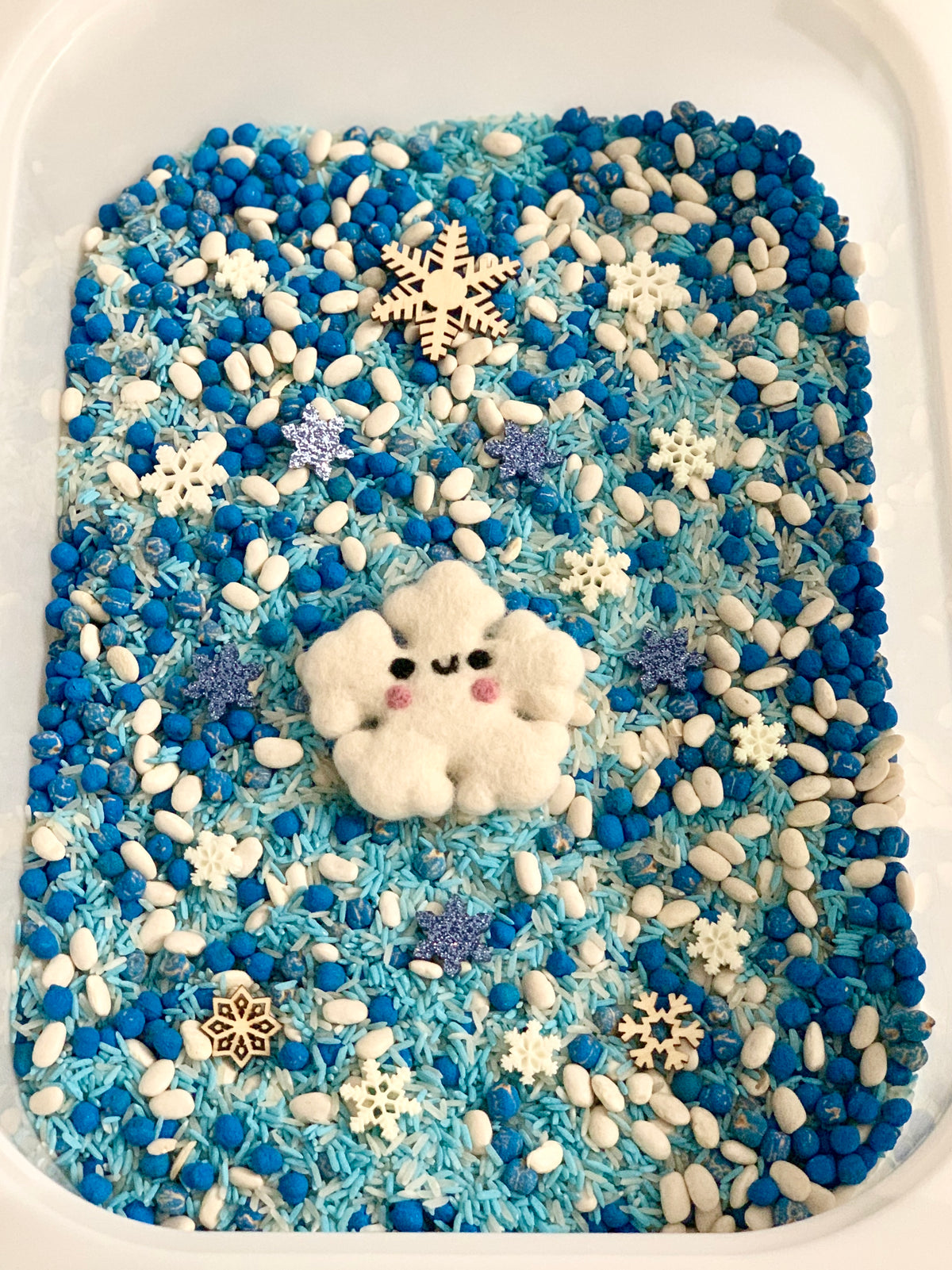 Blue Christmas sensory bin filled with blue and white rice, beans, snowflake shapes, and a smiling felt snowflake centerpiece