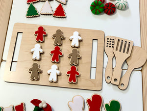 Wooden cookie sheet with red, white, and brown gingerbread cookies and wooden kitchen utensils on white table