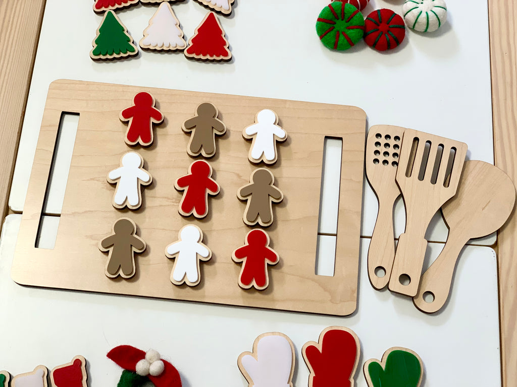 Wooden cookie sheet with red, white, and brown gingerbread cookies and wooden kitchen utensils on white table
