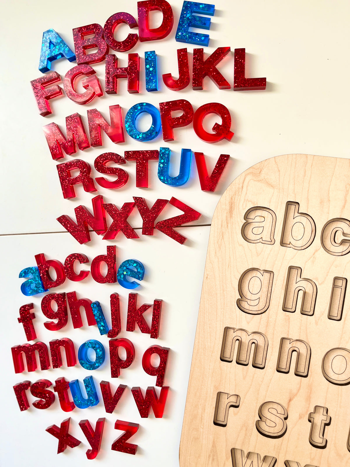 Red and blue glittery resin uppercase and lowercase alphabet letters arranged next to a wooden letter puzzle board