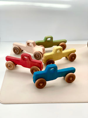 Five handcrafted wooden toy pickup trucks painted in blue, red, yellow, green, and natural wood on a light surface