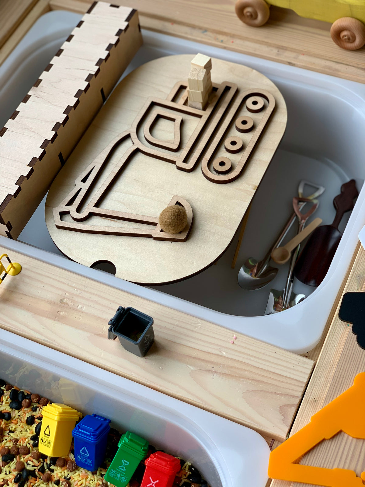 Wooden excavator puzzle piece placed inside a white container surrounded by small toy tools and colorful toy recycling bins nearby