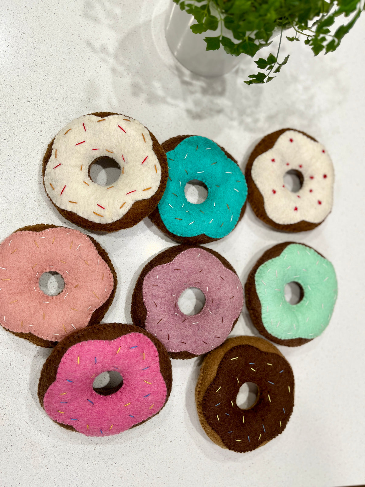 Eight felt donuts with colorful frosting and stitched sprinkles arranged on a white surface near a green potted plant