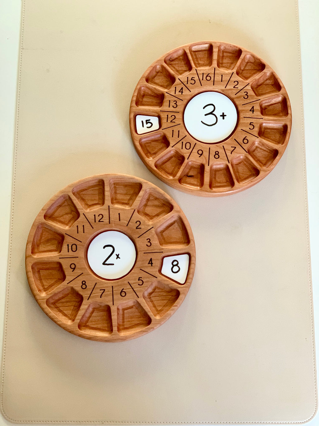 Two round wooden game boards with numbered triangular compartments and central multiplication symbols on a beige surface